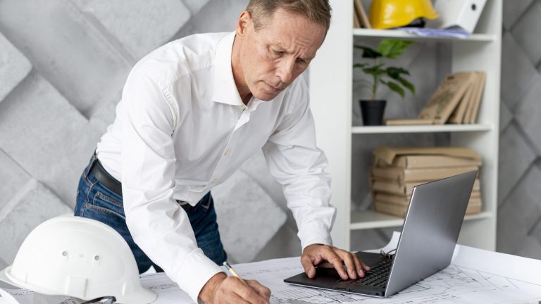 Man workin on blueprints with laptop in front of him and hardhat by his side illustrates blog "What Are the Most Important Project Management Tools?"