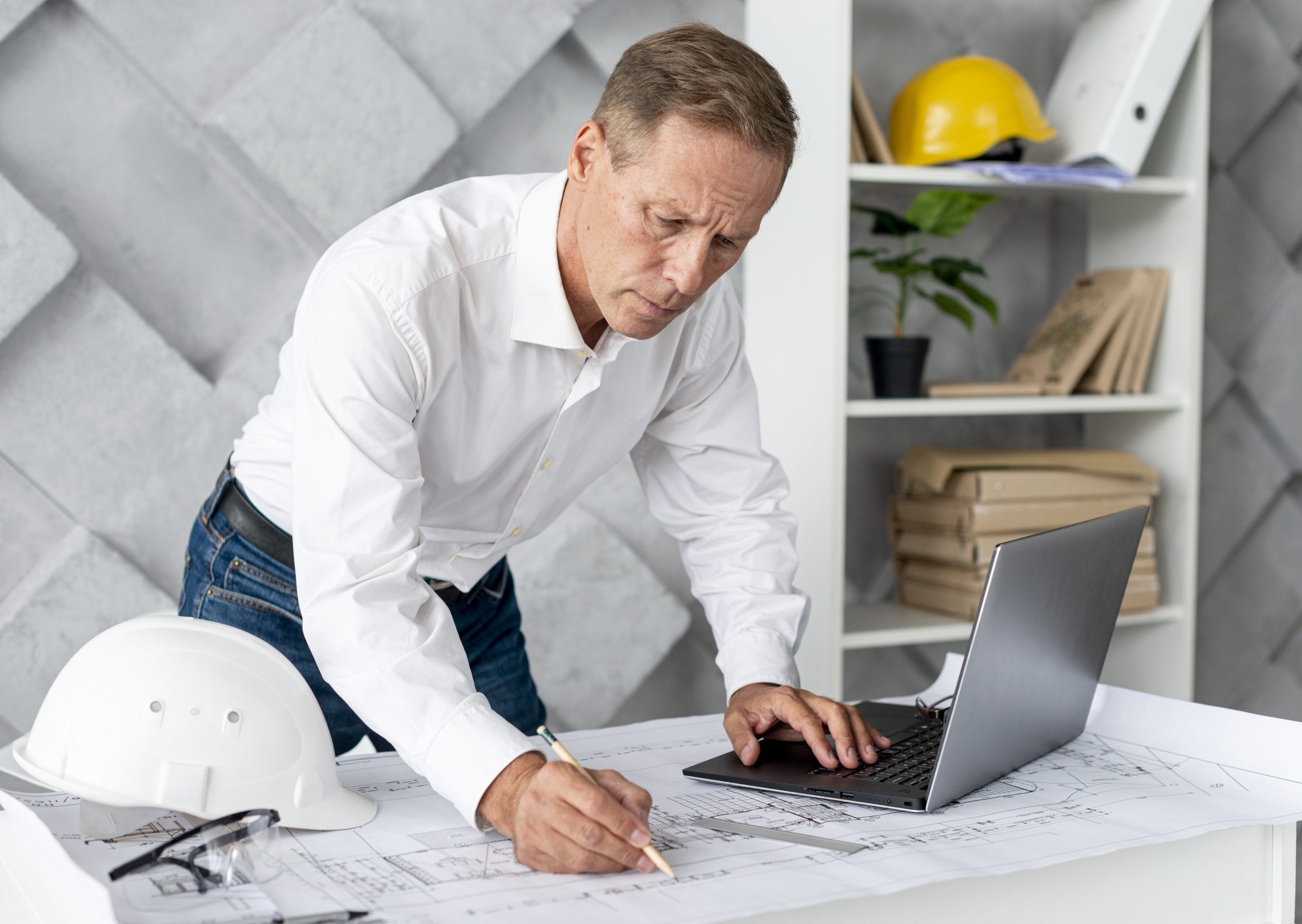 Man workin on blueprints with laptop in front of him and hardhat by his side illustrates blog "What Are the Most Important Project Management Tools?"