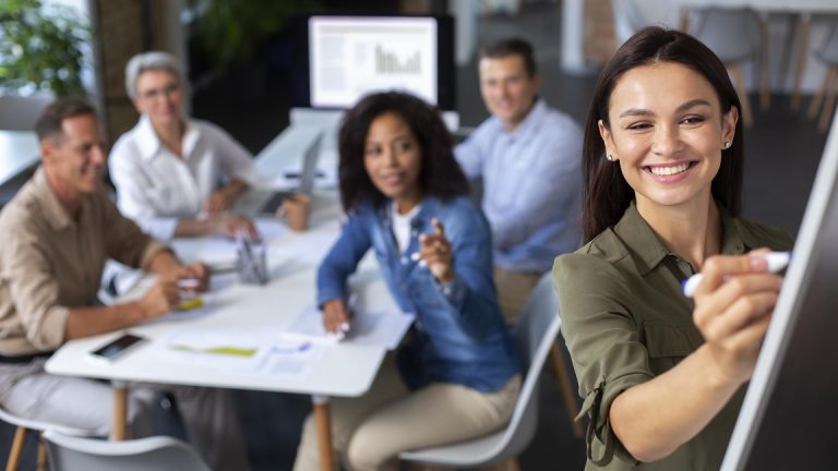 Woman writing on whiteboard with people in the background.